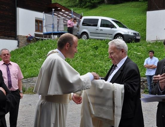 Ein Priester in einer weißen Robe übergibt einem älteren Mann in einem schwarzen Anzug ein weißes Tuch, mit Zuschauern und einem Van im Hintergrund.