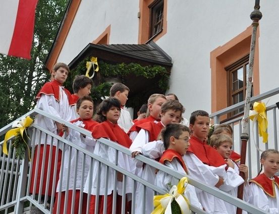 Eine Gruppe von Kindern in roten und weißen Gewändern steht auf einer Treppe, wahrscheinlich für eine Zeremonie, mit einer Flagge und Dekorationen im Hintergrund.
