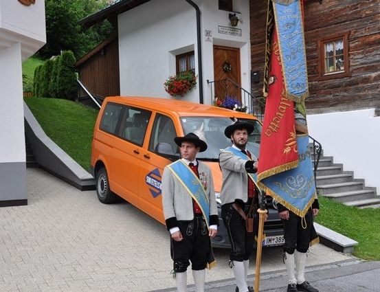 Drei Männer in traditioneller Kleidung stehen vor einem Haus mit einem geparkten Van davor. Der Mann auf der linken Seite trägt eine Schärpe und einen Hut, während die anderen eine Fahne halten.
