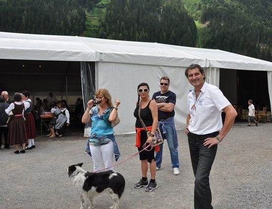 Eine Gruppe von Menschen, darunter zwei Frauen mit einem Hund, stehen vor einem weißen Zelt mit Bergen im Hintergrund.