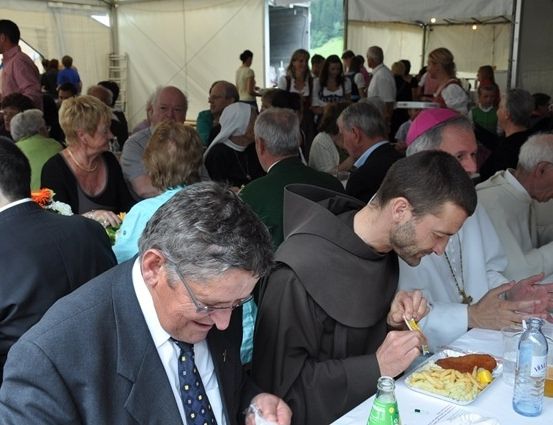 Eine Gruppe von Menschen, darunter ein Priester und ein Mönch, sitzen an einem Tisch in einem Zelt und essen und trinken.