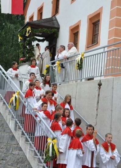 Eine Gruppe von Kindern in roten und weißen Gewändern steigt die Stufen einer Kirche hinauf, begleitet von Erwachsenen in weißen Gewändern. Die Kirche ist mit gelben Bändern und Grünpflanzen geschmückt.