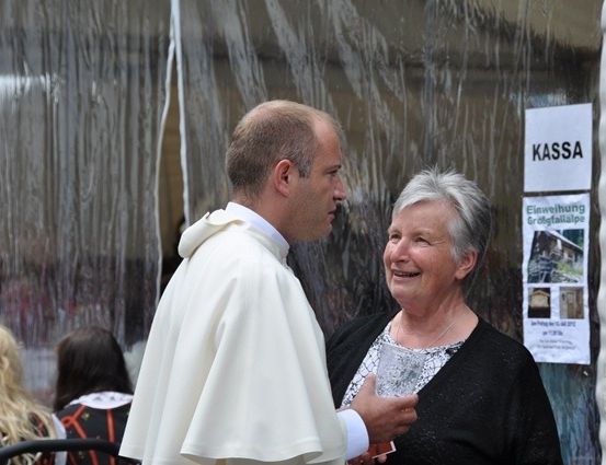 Ein Priester in einer weißen Robe unterhält sich mit einer älteren Frau in einem schwarzen Pullover, die vor einer Glastür mit einem Schild 'Kassa' stehen. Im Hintergrund sind verschwommen Menschen und ein weißer Vorhang zu sehen.