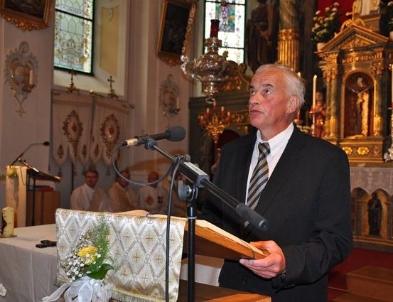 Ein Mann in einem Anzug steht an einem Pult in einer Kirche und liest aus einem Buch. Hinter ihm stehen ein Mikrofon und Blumen auf dem Pult. Im Hintergrund sind ein Priester und ein Altar zu sehen.