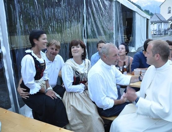 Eine Gruppe von Menschen, einschließlich eines Priesters, sitzen um einen Tisch herum, lächeln und unterhalten sich. Sie sind in traditioneller Kleidung gekleidet.