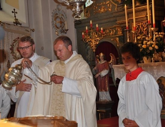 Ein Priester hält einen silbernen Kelch, während ein anderer Priester und zwei Messdiener hinter ihm in einer Kirche stehen.