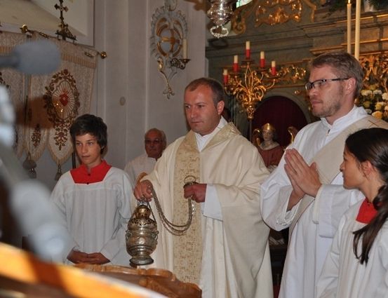 Ein Priester, der eine Kerze hält, steht vor einem Altar mit einem Mikrofon, während zwei andere Männer und zwei Frauen in weißen Gewändern hinter ihm stehen.