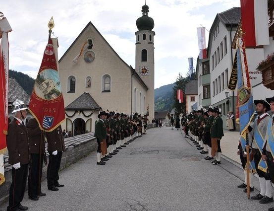 Ein zeremonieller Aufmarsch uniformierter Personen steht vor einer Kirche mit Fahnen und einem Glockenturm an einem bewölkten Tag.