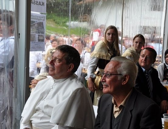 Zwei Männer in Anzügen sitzen an einem Tisch mit einem Priester in einer weißen Robe, während eine Frau in traditioneller Kleidung hinter ihnen steht. Die Szene spielt sich in einem Raum mit Glaswänden ab, vor dem eine Menschenmenge steht.