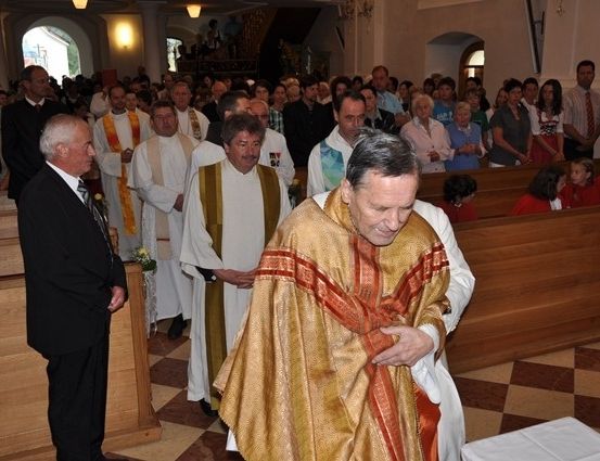 Ein Priester in einer goldenen Robe wäscht die Füße eines anderen Mannes in einer Kirche. Viele Menschen stehen und sitzen im Hintergrund.