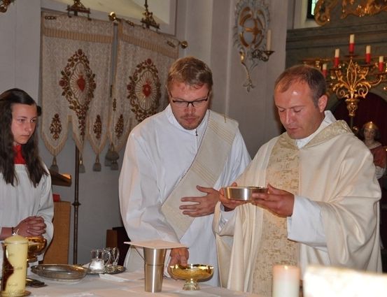 Zwei Priester in weißen Gewändern führen einen Ritus in einer Kirche durch, mit Kerzen und einem Kelch auf dem Tisch vor ihnen.