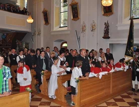 Eine Gruppe von Menschen, darunter Kinder in weißen Kleidern, steht während einer Zeremonie in einer Kirche. Einige halten Blumen, und ein Mann in grüner Uniform steht vorne.