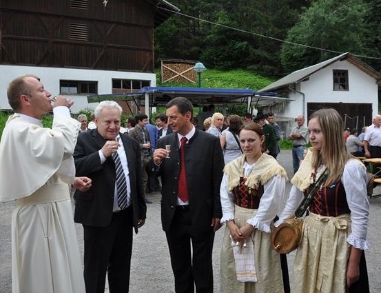 Ein Priester trinkt aus einem Glas, während er mit zwei Männern und zwei Frauen in traditioneller Kleidung vor einem Gebäude steht.