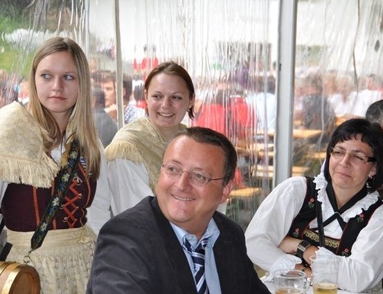 Ein Mann in Anzug und drei Frauen in traditioneller Kleidung sitzen an einem Tisch in einem überfüllten Restaurant. Der Mann lächelt, während die Frauen in verschiedene Richtungen schauen. Dahinter sitzen verschwommene Menschen an anderen Tischen.