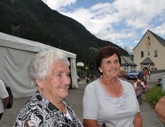 Zwei ältere Frauen stehen lächelnd vor einer Kirche, mit einem Berg und einem Zelt im Hintergrund.