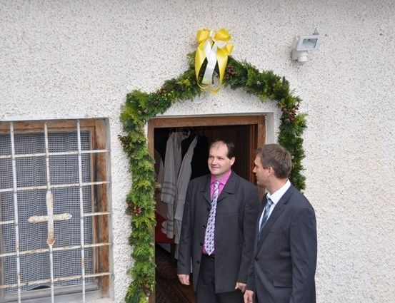 Zwei Männer in Anzügen stehen vor einem Gebäude mit einer Weihnachtsgirlande und einem Kreuz am Fenster.