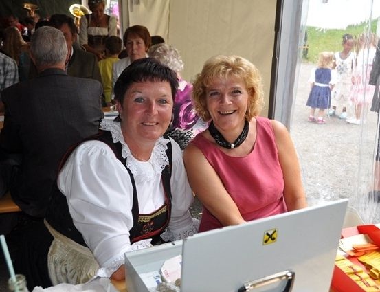 Zwei Frauen sitzen an einem Tisch mit einem Laptop vor ihnen. Eine trägt ein traditionelles Kleid, die andere ein rosa Oberteil. Dahinter sitzen eine Gruppe von Menschen an Tischen, und ein paar Kinder stehen draußen.