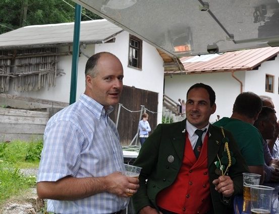 Zwei Männer, einer in einer traditionellen grünen und roten Kleidung, stehen unter einem Vordach und halten Getränke. Hinter ihnen sitzen eine Gruppe von Menschen an einem Tisch. Im Hintergrund ist ein Haus mit einem roten Dach und einer Treppe zu sehen.