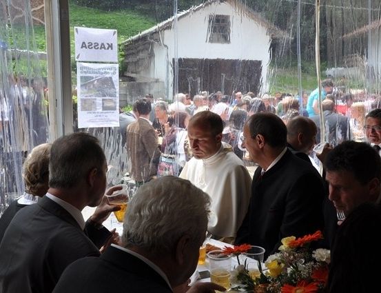 Eine Gruppe von Männern in Anzügen sitzt um einen Tisch mit Blumen und Getränken, während ein Priester in Weiß in der Nähe steht. Eine Menschenmenge hat sich draußen versammelt, und es gibt ein Schild am Fenster.