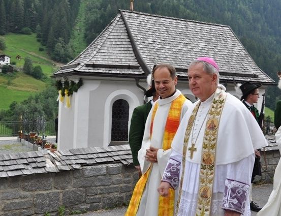 Zwei Priester in weißen Gewändern stehen vor einer kleinen Kapelle mit einer Steinmauer und einem grünen Feld im Hintergrund.