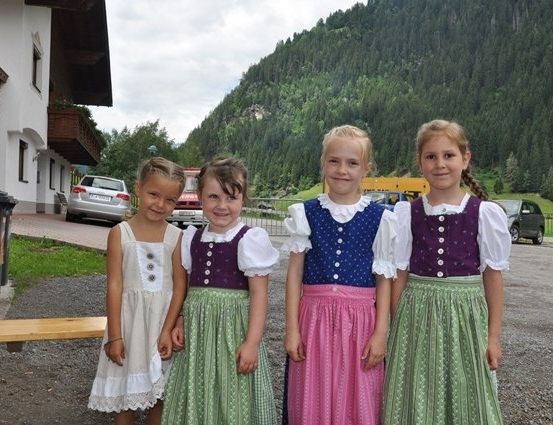 Vier junge Mädchen in traditioneller Kleidung stehen vor einem Gebäude mit einem Berg im Hintergrund.