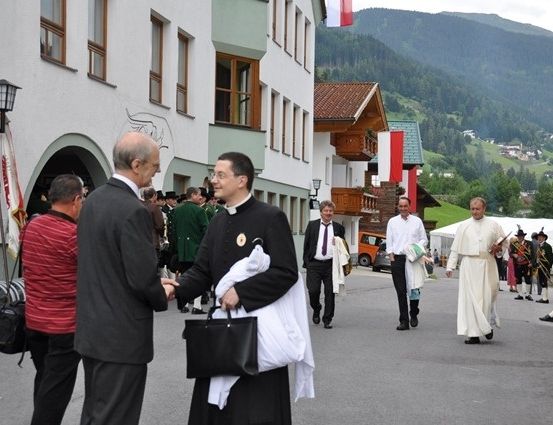 Eine Gruppe von Menschen, einschließlich Geistlicher, geht auf einer Straße vor einem Gebäude mit Fahnen und Bergen im Hintergrund.