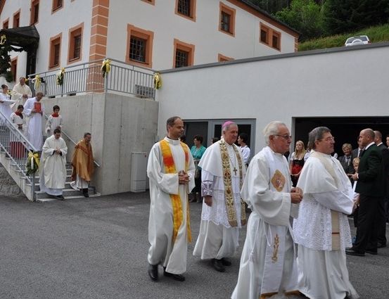 Mehrere Priester in weißen Gewändern und Stola gehen in einer Prozession vor einem Kirchengebäude. Einige Leute stehen auf den Stufen und andere beobachten.
