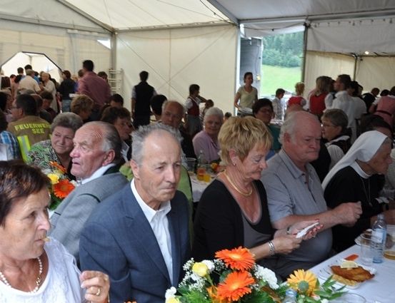Eine Gruppe älterer Menschen sitzt an einem langen Tisch in einem Zelt, mit Blumen und Essen auf dem Tisch. Sie sind in formeller Kleidung gekleidet.