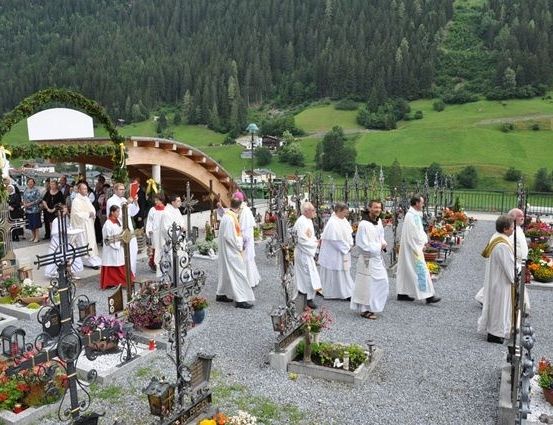 Eine Gruppe von Priestern in weißen Gewändern versammelt sich um einen Friedhof mit Blumen und Kreuzen in einem Gebirgsgebiet.