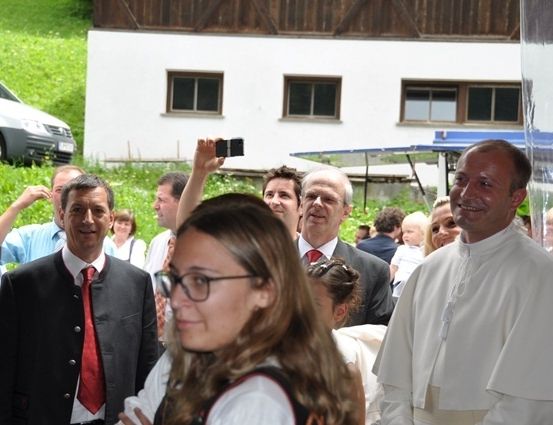 Eine Gruppe von Menschen, darunter ein Mann in einem roten Schal, versammelt sich vor einem Gebäude. Ein Mann in einem weißen Gewand lächelt und macht ein Selfie mit einem Handy.