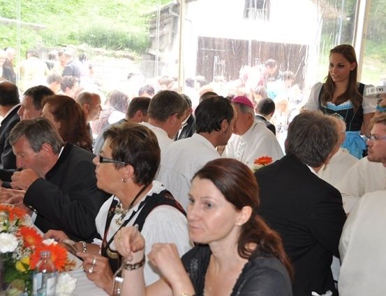 Eine Gruppe von Menschen sitzt an einem langen Tisch in einem Restaurant. Einige tragen Brillen und Halsketten. Eine Frau steht hinter dem Tisch und hält eine Tablett. Es gibt Blumen auf dem Tisch. In der Ferne stehen Menschen und beobachten.