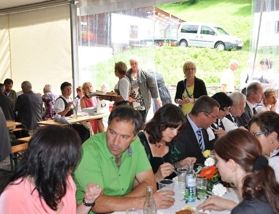 Eine Gruppe von Menschen isst in einem Zeltrestaurant im Freien. Ein Mann in einem grünen Hemd sitzt an einem Tisch mit einer Frau in einem rosa Hemd. Eine Frau in einer schwarzen Jacke geht mit einem Tablett. Ein weißer Van ist draußen geparkt.