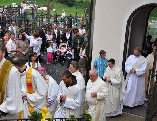 Eine Gruppe von Priestern in weißen Gewändern steht vor einer Kirche, mit einer Menge von Menschen hinter ihnen.