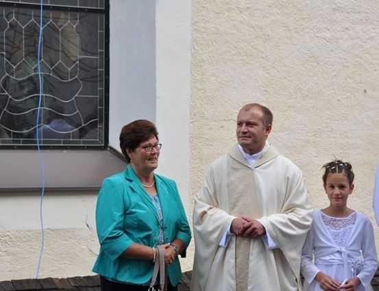 Ein Priester steht neben einer Frau und einem jungen Mädchen, alle lächelnd, vor einer Kirche.