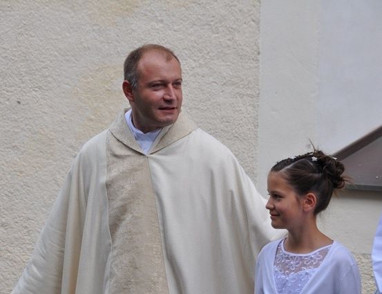 Ein Priester in einer weißen Robe steht neben einem jungen Mädchen in einem weißen Kleid, beide lächeln und schauen in die Kamera.