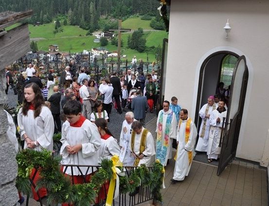 Eine Gruppe von Menschen in weißen Gewändern versammelt sich vor einer Kirche, einige betreten sie durch die offene Tür. Die Kirche ist von Grün und Bergen im Hintergrund umgeben.