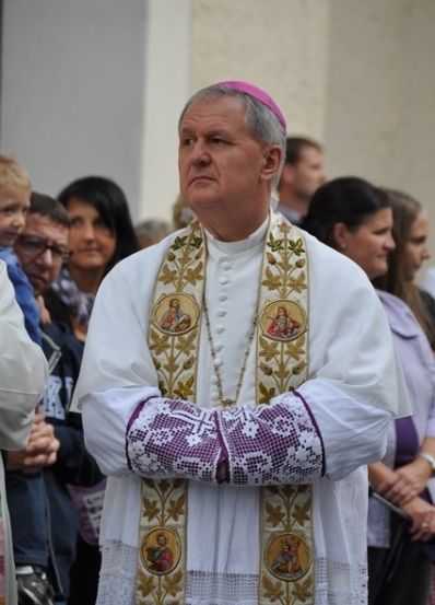 Ein Priester in einer weißen Robe und einer rosa Biretta steht vor einer Menschenmenge, möglicherweise während einer religiösen Zeremonie.