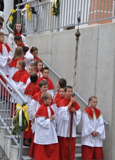Eine Gruppe von Kindern in roten und weißen Gewändern steigt die Stufen einer Kirche hinauf und hält einen Stab.