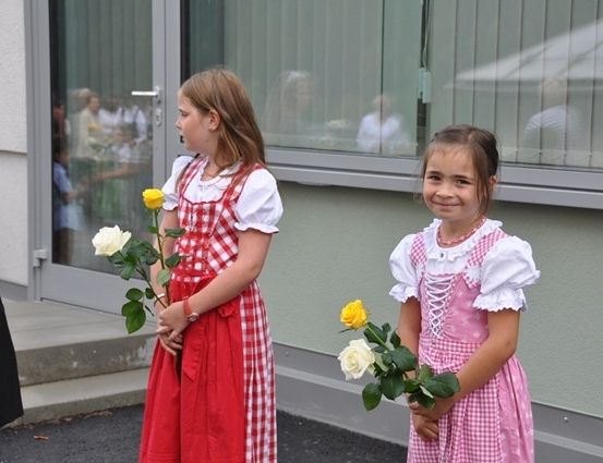 Zwei junge Mädchen in traditionellen bayerischen Kleidern halten gelbe und weiße Rosen vor einem Gebäude mit großen Fenstern.