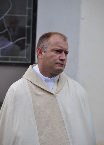 Ein Priester in einer weißen Robe steht vor einem Gebäude und schaut nach links. Das Gebäude hat ein Glasfenster mit einem geometrischen Muster.
