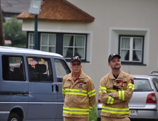Zwei Feuerwehrleute stehen vor einem Haus, vor dem ein blauer Van und ein silberner Wagen geparkt sind.