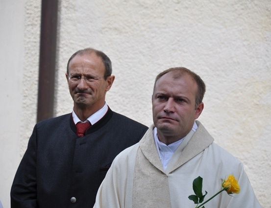 Zwei Männer stehen vor einer weißen Wand; einer trägt eine rote Krawatte und eine Brille, der andere hält eine gelbe Rose.