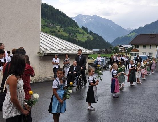Eine Gruppe von Menschen, darunter Kinder mit Blumen, steht in einer Reihe auf einer nassen Straße. Ein Mann im Rollstuhl ist unter ihnen. Hinter ihnen befinden sich Gebäude und eine Berglandschaft.