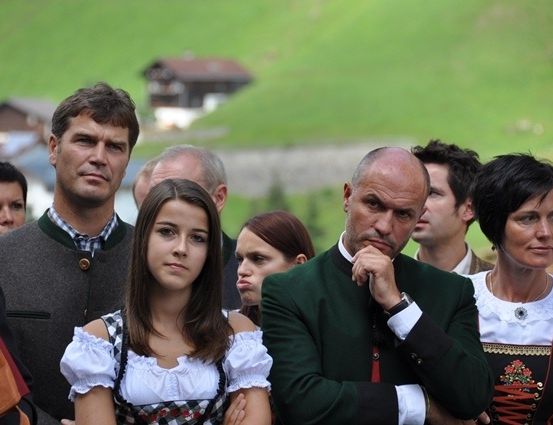Eine Gruppe von Menschen in traditioneller Kleidung steht in einer Reihe, wahrscheinlich bei einem Festival, mit einer malerischen Bergkulisse im Hintergrund.