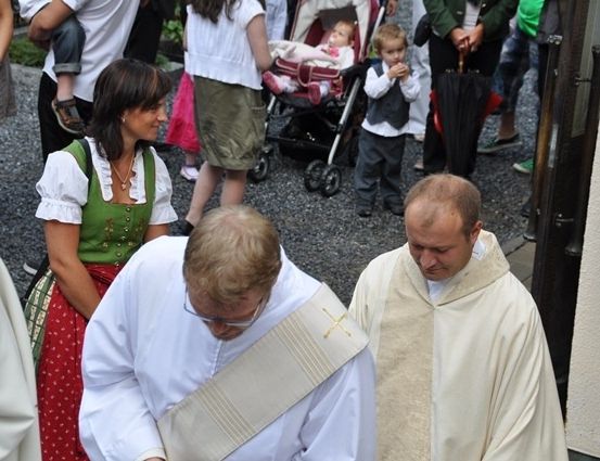 Zwei Priester in weißen Gewändern gehen zusammen, einer von ihnen verneigt sich. Eine Frau in einem grünen Kleid und ein Mann in einem weißen Hemd stehen hinter ihnen. Im Hintergrund sind Menschen zu sehen, darunter ein Baby in einem Kinderwagen.