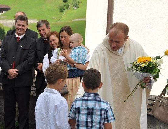 Ein Priester hält Blumen und steht mit Kindern und Erwachsenen, möglicherweise bei einer Kirchenveranstaltung.