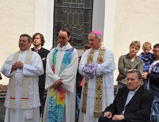 Eine Gruppe von Männern in weißen Gewändern, darunter zwei Priester, stehen vor einer Kirche mit einem Buntglasfenster. Ein Mann im Rollstuhl sitzt vor ihnen.