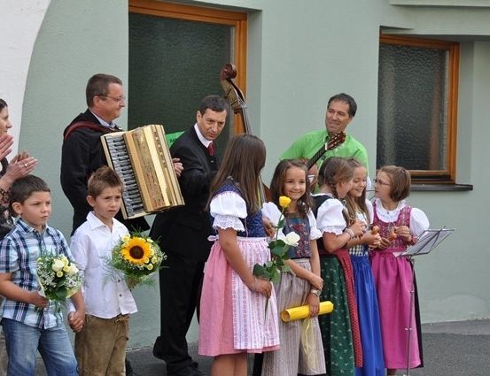 Eine Gruppe von Kindern und Erwachsenen steht vor einem Gebäude, einige halten Blumen und Musikinstrumente.