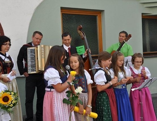 Eine Gruppe von Kindern in traditioneller Kleidung, die Blumen und Musikinstrumente halten, steht vor einem Gebäude, hinter dem Musiker spielen.