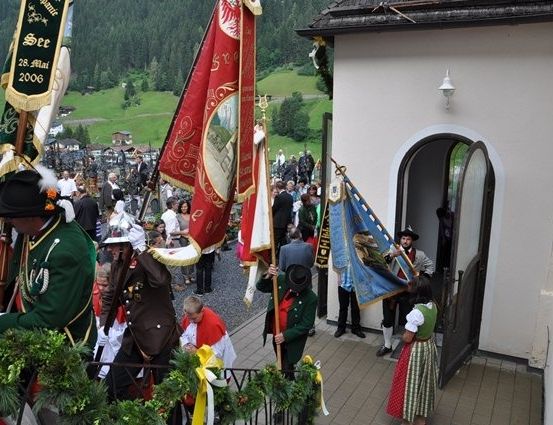 Eine Gruppe von Menschen in traditioneller Kleidung steht vor einer Kirche mit Fahnen und Bannern, möglicherweise während eines Festivals.